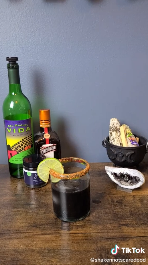 Spooky margarita is black and garnished with Tajin rim and slice of lime. Del Maguey Vida mezcal bottle and Cointreau bottle are displayed with activated charcoal jar. Cauldron with sage in front of stone in background.