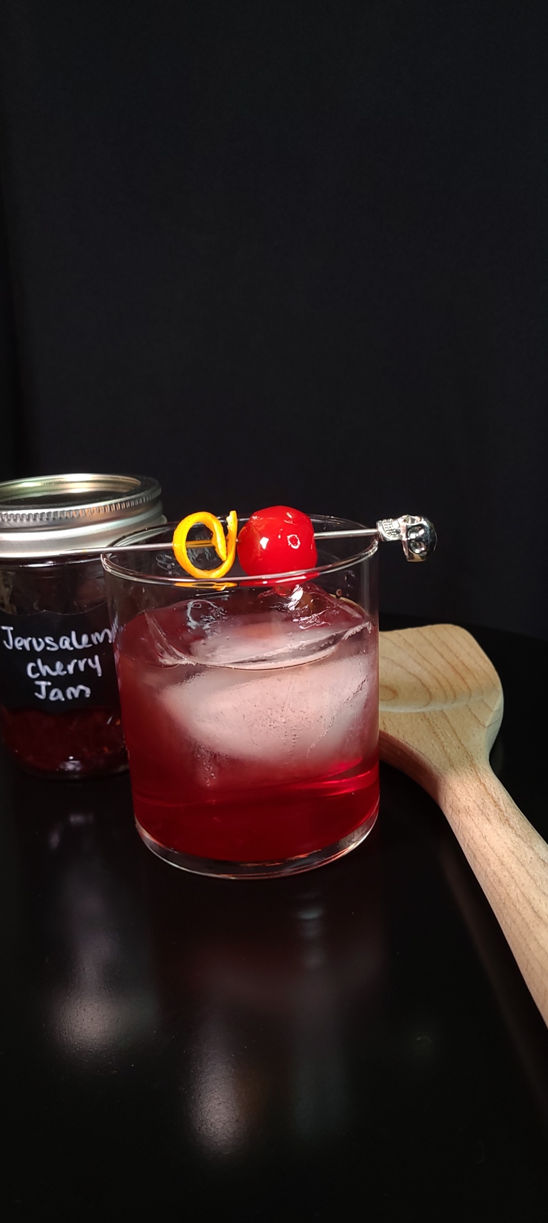 The Old Fashioned Gratitude cocktail sits on a black table with a wooden spoon and a jar labeled "Jerusalem Cherry Jam". The drink is red and garnished with a maraschino cherry and orange peel.
