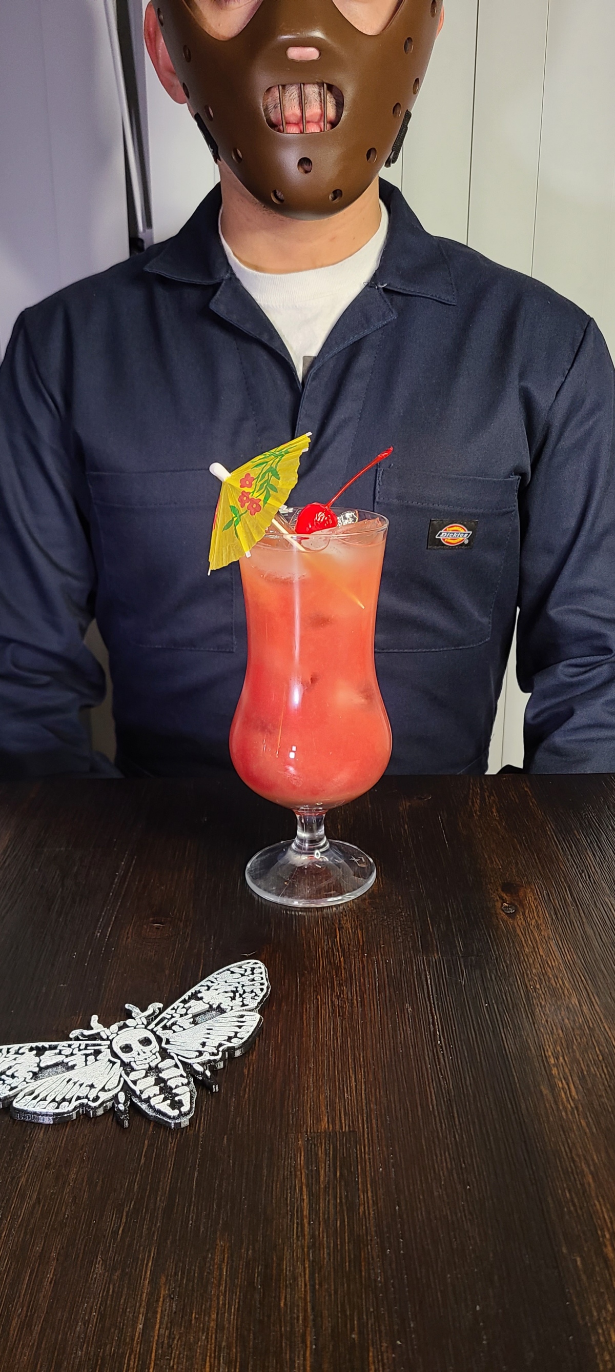 The Hannibal on the Beach drink sits on a dark wood table with a 3D print of the Silence of the Lambs moth. The drink is a reddish pink and is garnished with an umbrella and cherry.